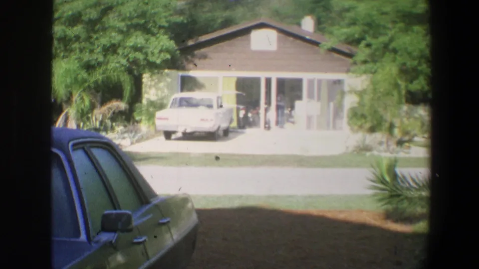 1973 Florida home-movie view of a single-story house, driveway vehicle, palms, and a gray car partly blocking the foreground.
