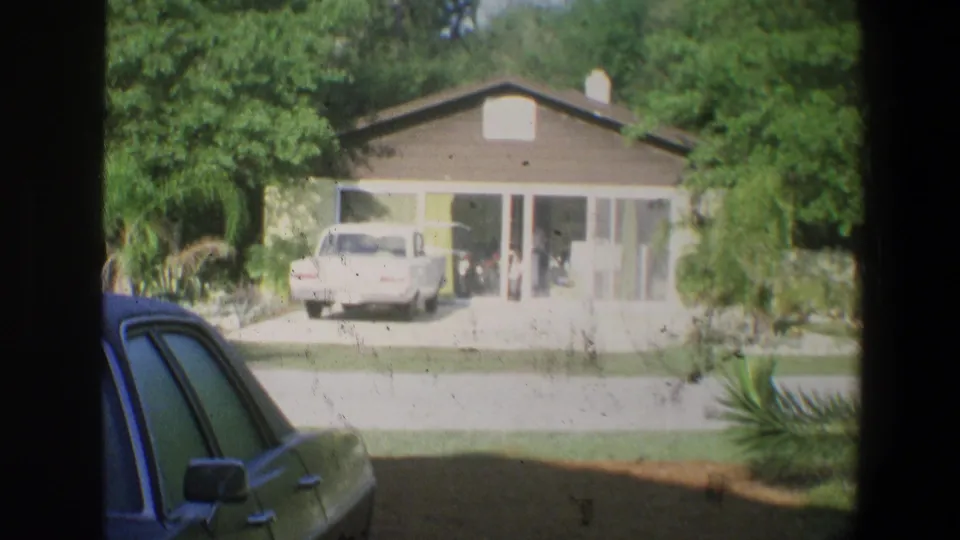 1973 8mm Florida residential view with a white vehicle in a driveway, broad house frontage, greenery, and a gray car at frame edge.