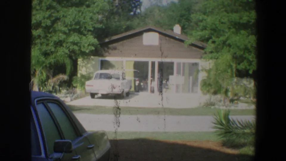1973 Florida 8mm frame showing a sunlit house, driveway vehicle, gray car in foreground, palms, and small distant figures near the entry.