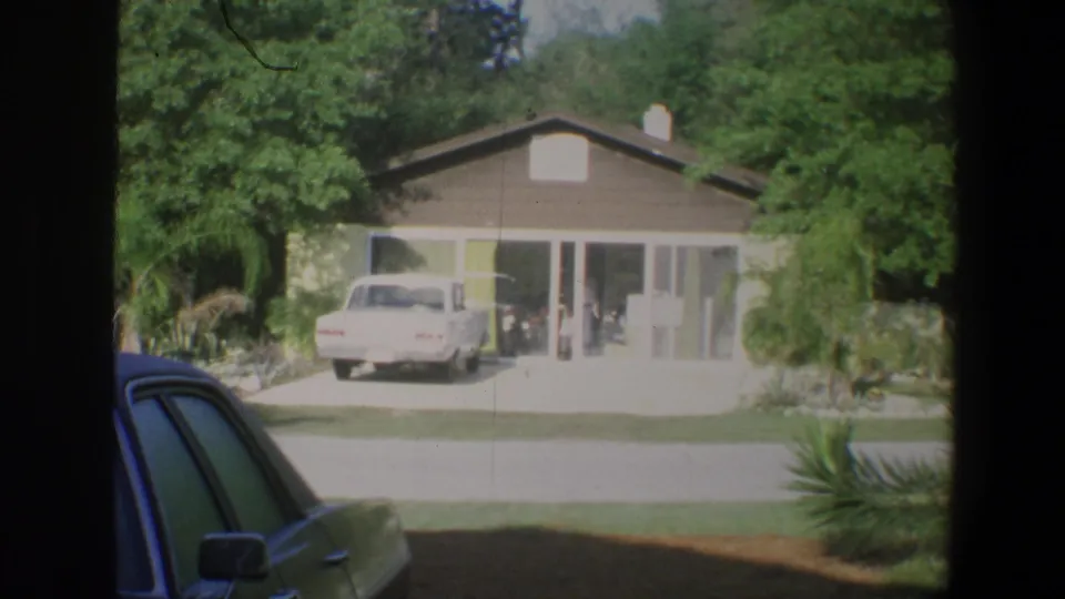 1973 Walt Disney World Florida home-movie frame showing a bungalow, driveway, white car, trees, and dark car-window edges.