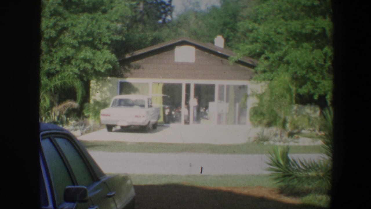 1973 Walt Disney World home-movie frame showing a low modern building, driveway, white car, trees, and palm plantings.
