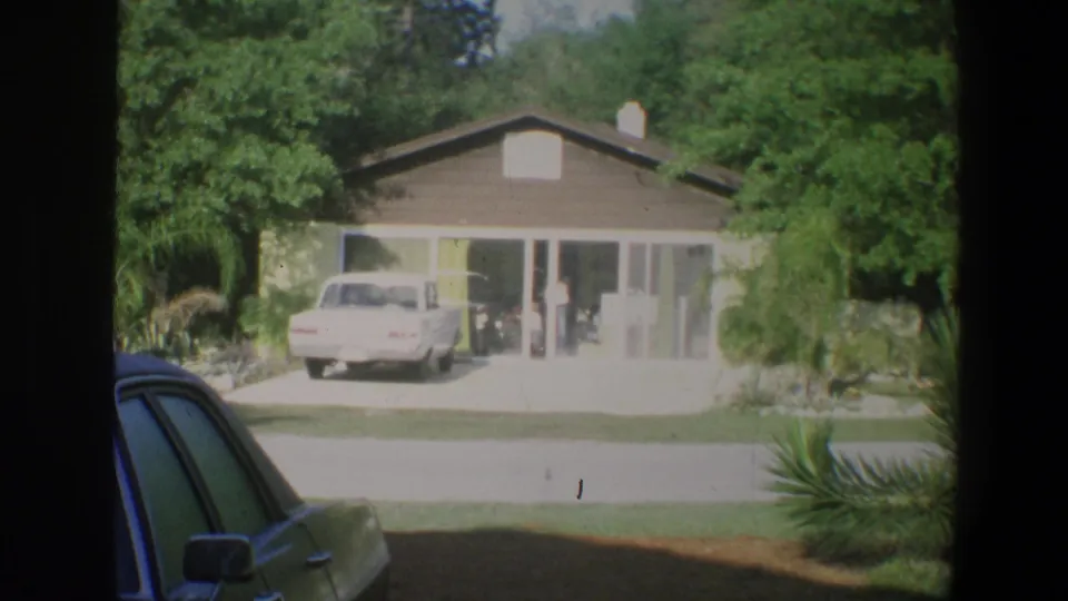1973 Walt Disney World home-movie frame showing a low modern building, driveway, white car, trees, and palm plantings.