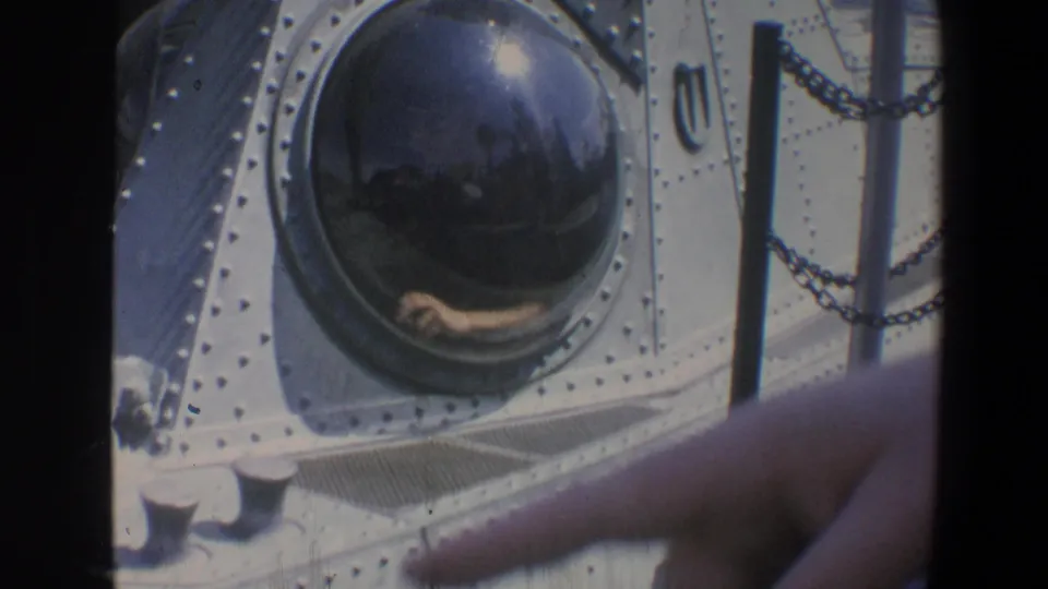 1973 Walt Disney World home-movie frame of a large submarine porthole on riveted panels, partly obscured by the camera operator's finger.
