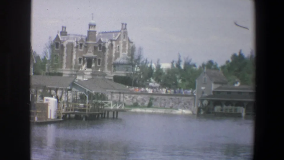Across the water at Walt Disney World in 1973, a castle-like building rises behind docks, low walls, trees, and distant visitors.