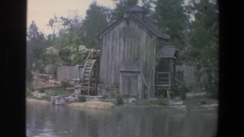 A weathered wooden mill and waterwheel stand along the Walt Disney World shoreline in 1973, surrounded by trees and calm water.