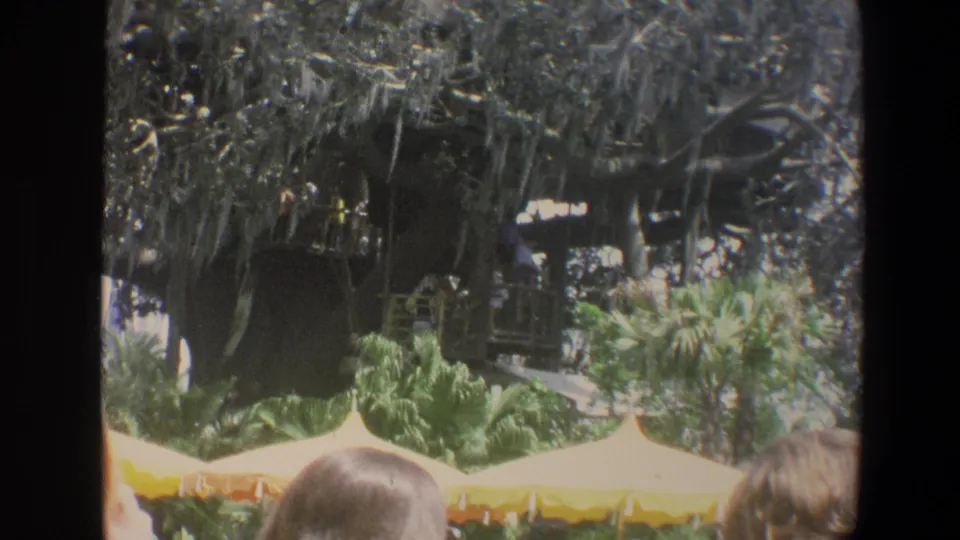 1973 Walt Disney World home-movie frame showing a thatched structure, mossy tree, yellow canopy, tropical plants, and partial guests.