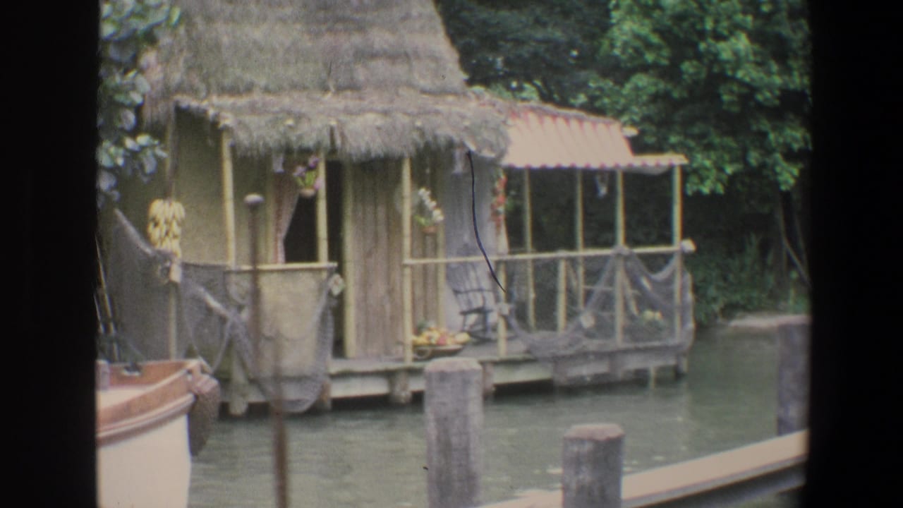 Thatched hut beside a canal with nets, hanging fruit, and a boat edge in the foreground.