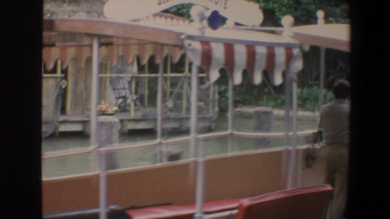 Striped awning boat passes a canal-side hut with a man standing near the helm.