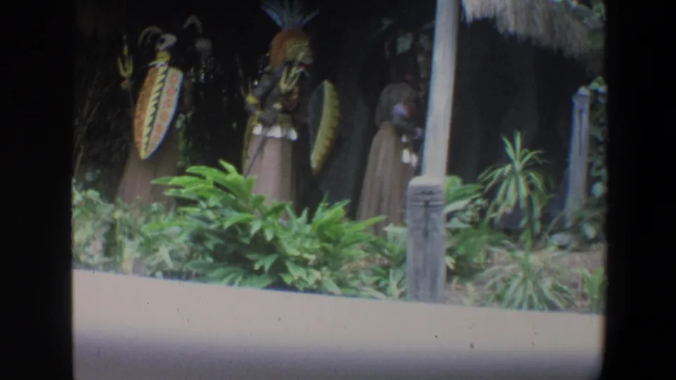 1973 Walt Disney World 8mm frame showing tribal-themed attraction figures in a dark landscaped setting with plants and a thatched edge.