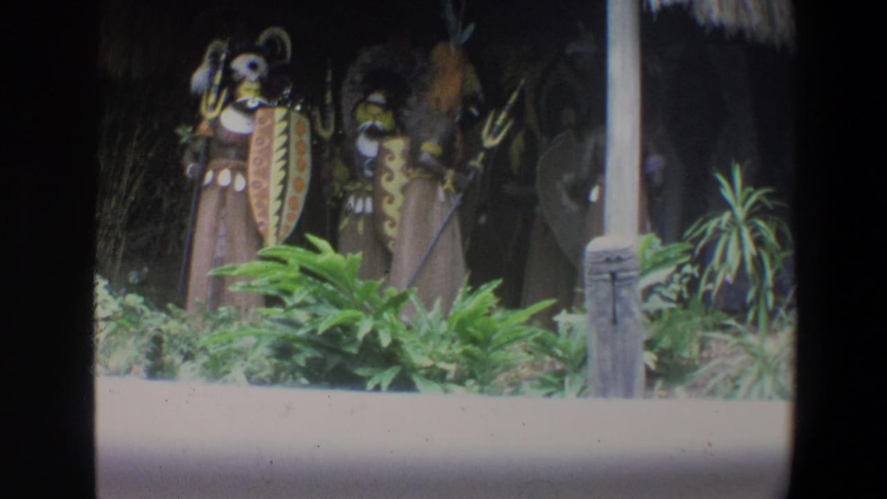 1973 Walt Disney World 8mm frame showing blue and orange costumed display figures near shields, posts and dense greenery.