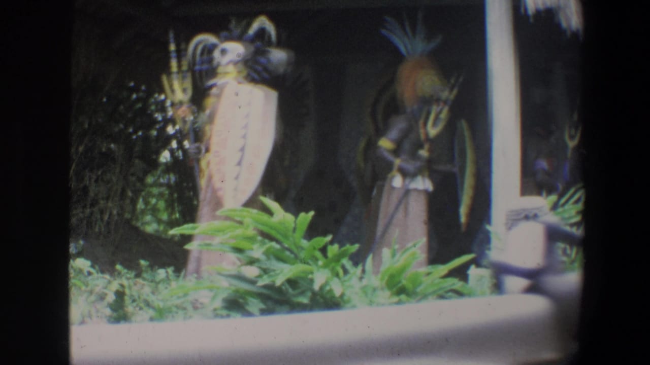 Two costumed display figures stand inside a dark hut with shields, props and foreground plants.