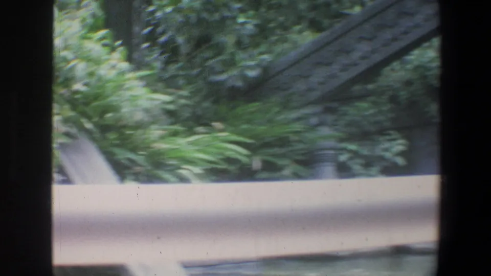 1973 Walt Disney World home-movie frame showing a pale railing, tropical foliage, and a stone structure during a handheld walk.