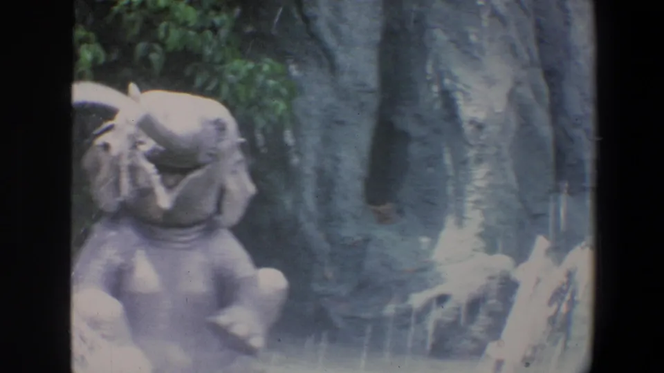 1973 Walt Disney World home-movie frame showing an elephant figure with its trunk raised near rockwork and foliage.