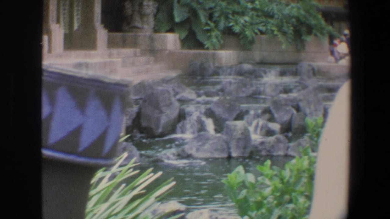 1973 Walt Disney World home-movie frame showing a tropical-themed thatched building, rock waterfall, pond, plants, and a blue patterned foreground object.