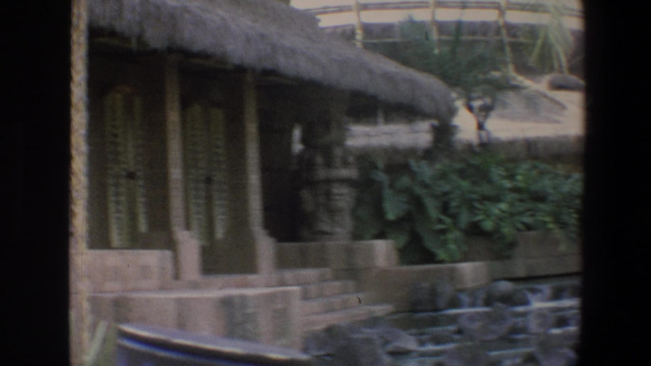 Thatched-roof building with posts, greenery, railings, and distant visitors near the upper walkway.
