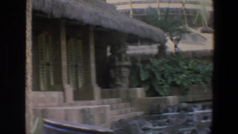 1973 Walt Disney World frame with thatched roofline, carved or wooden posts, planted greenery, railings, and small distant visitors in the background.