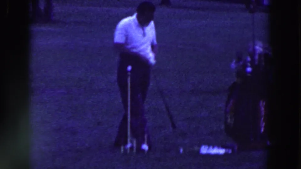 A man in a white polo and dark trousers stands with a golf club beside a bag on the grass in Ridgefield, New Jersey, 1961.