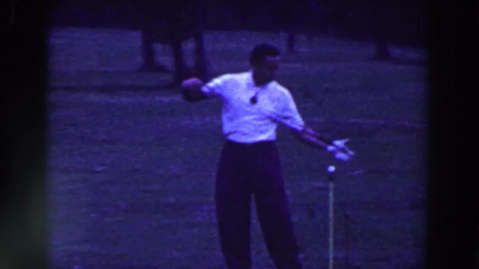 The golfer follows through on a shot in Ridgefield, New Jersey, with a teed ball, golf club, and shaded trees visible in the 1961 footage.
