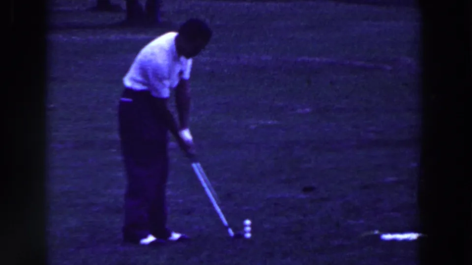The golfer bends over a ball or tee while holding the club low, part of a 1961 home-movie golf trick sequence in New Jersey.