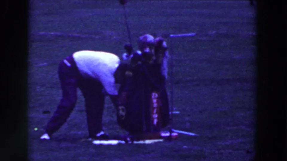 The golfer bends beside his bag and clubs, apparently adjusting equipment or the ball on the grassy course.