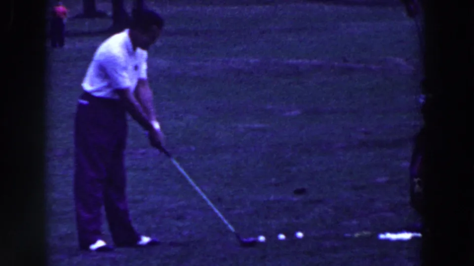 The golfer prepares to swing at a white ball on grass, with additional balls lined up nearby in the 1961 home-movie footage.