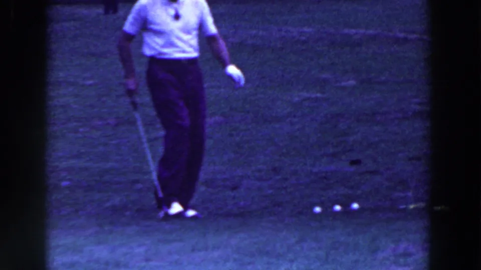 A man in golf attire walks across the grass holding his club, while several white balls remain visible near the lower edge.
