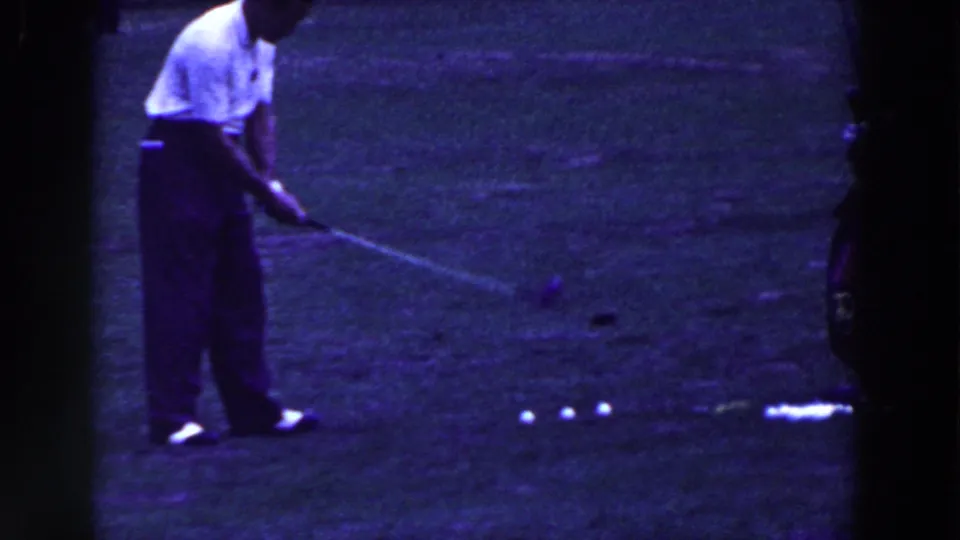 The golfer starts his swing in the purple-tinted 8mm frame, with three balls still lined on the grass.