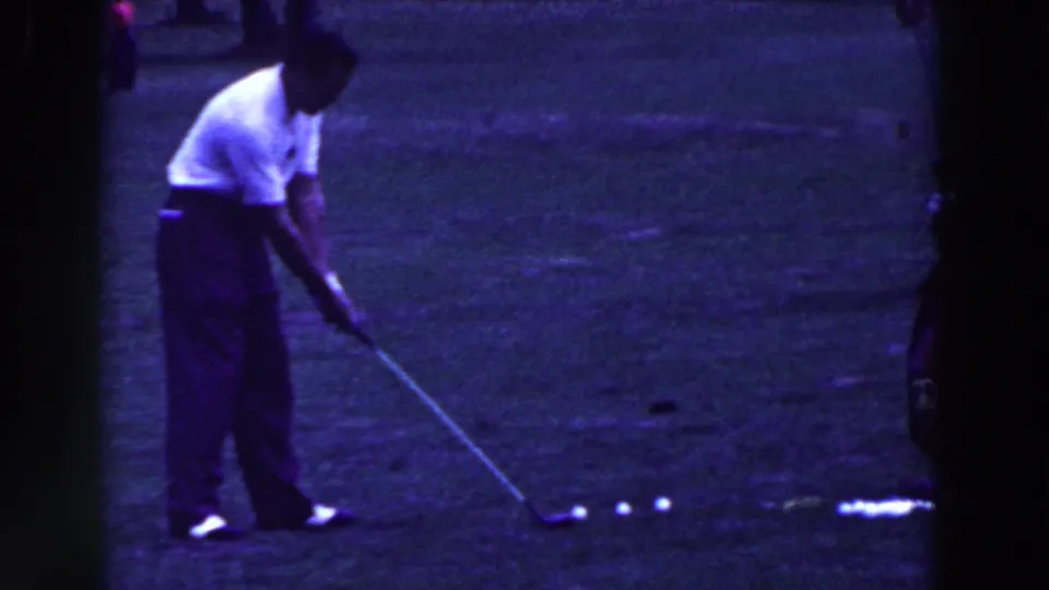 The golfer returns toward his address position, leaning over the club near the row of balls on the grass.