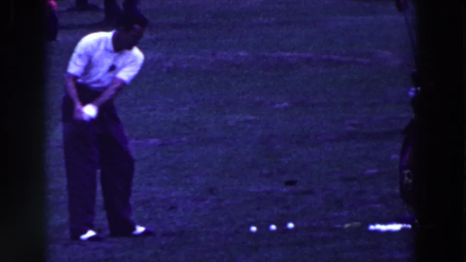 The man completes a golf swing motion in the 1961 home-movie footage, with balls visible to his side on the grass.