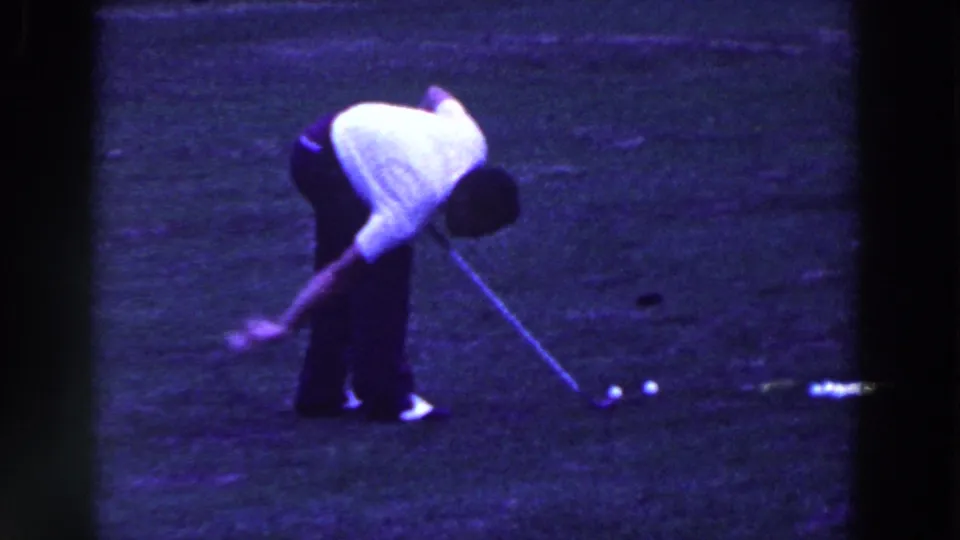 The golfer bends low with his club near two balls on the grass in vintage 8mm footage from Ridgefield, New Jersey.