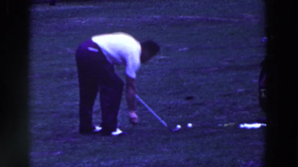 A man crouches over his golf club beside several balls on a grass course in 1961 Ridgefield home-movie footage.