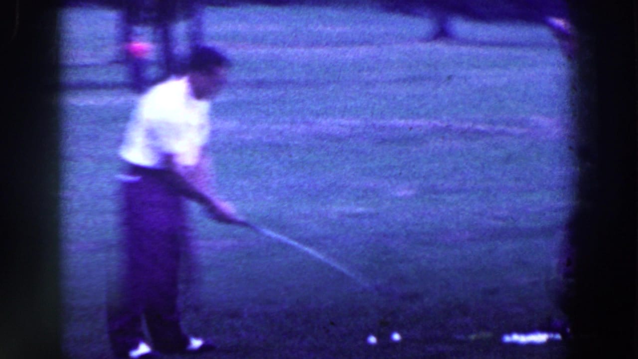 Man in a white shirt addresses a golf ball on a dark grassy course.