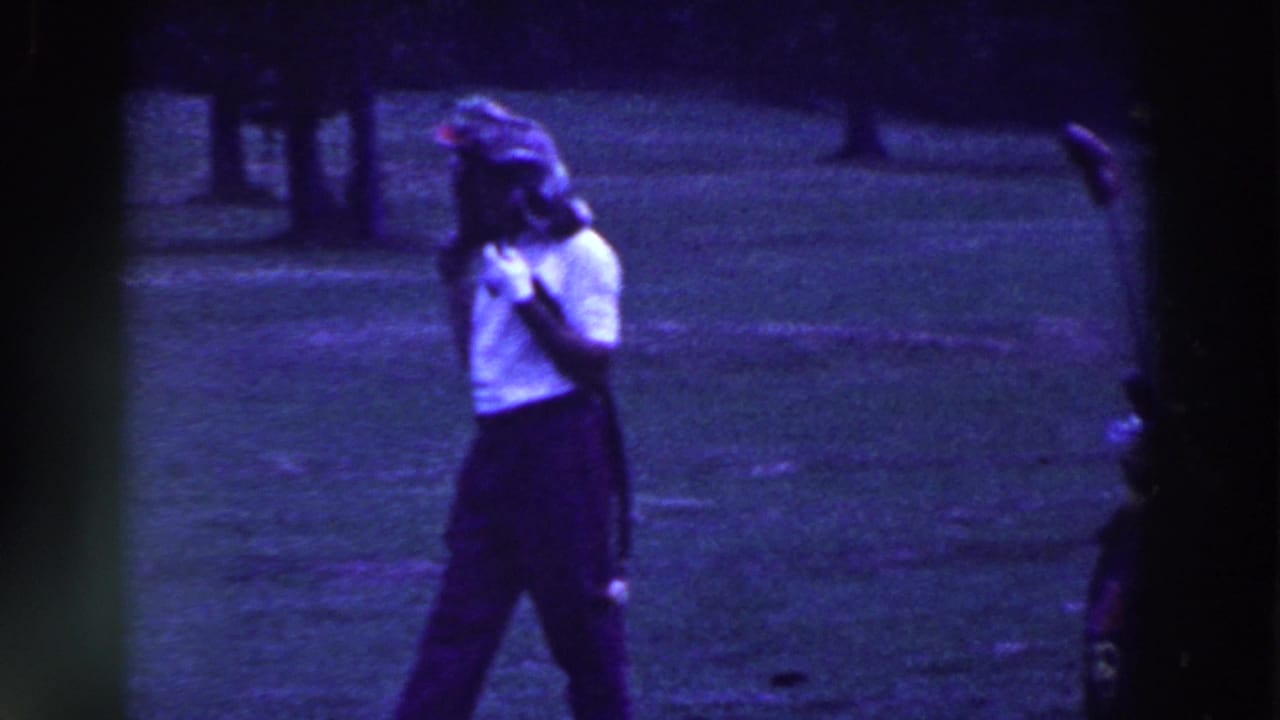Woman in a white blouse walks across a grassy golf course holding a golf club.