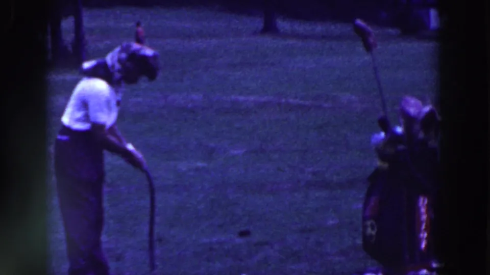 A man in dark pants bends over the putter near his golf bag during the 1961 Ridgefield golf scene.