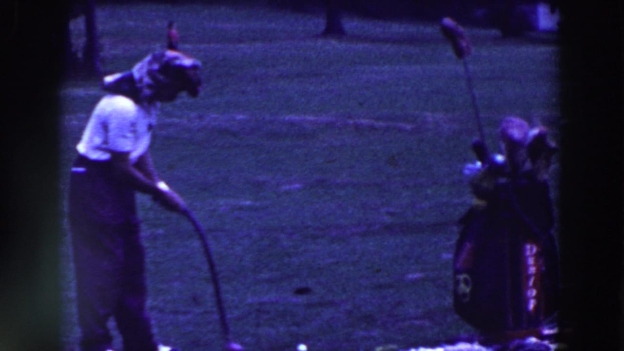 Man on grass bends toward a golf ball beside a golf bag with clubs.