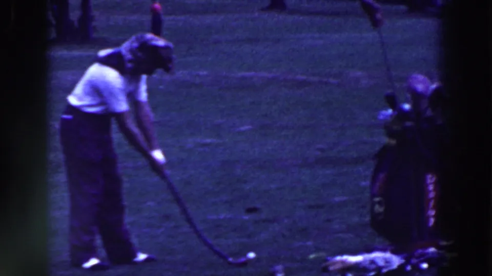 The golfer appears to guide a flexible club or prop toward a ball, with the golf bag and clubs visible at the edge of the frame.