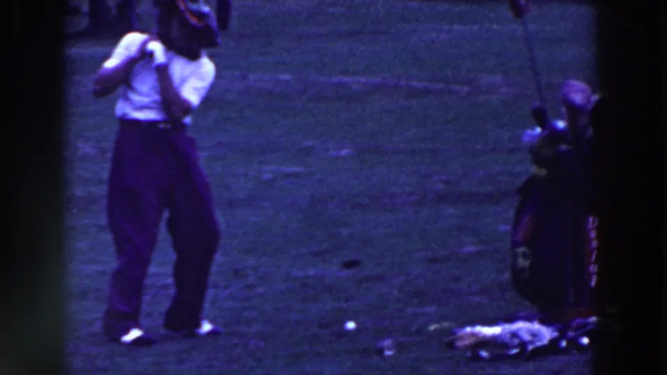 The man stands back from the ball after the attempt, with the club or prop raised and the golf bag still beside him on the course.