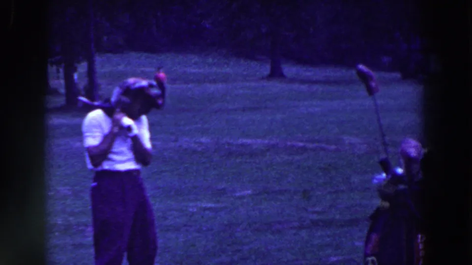 The golfer stands on the grass with the club or prop near his shoulder, while the golf bag and clubs remain visible in the 8mm frame.