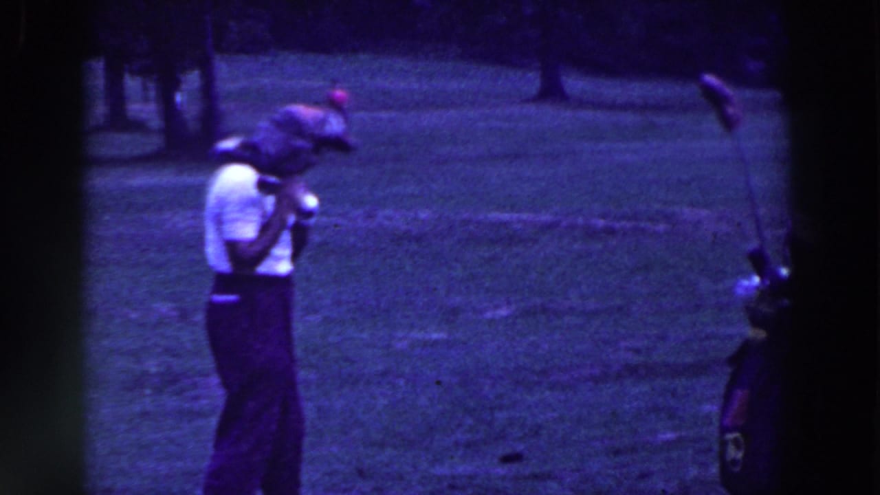 Man in white shirt raises a golf club near a golf bag on a grassy course.