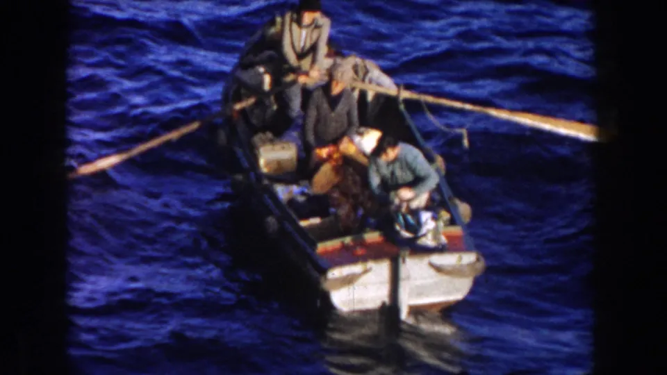 1959 Gibraltar home-movie frame showing an open boat with passengers, long oars, and luggage or bundles on vivid blue water.