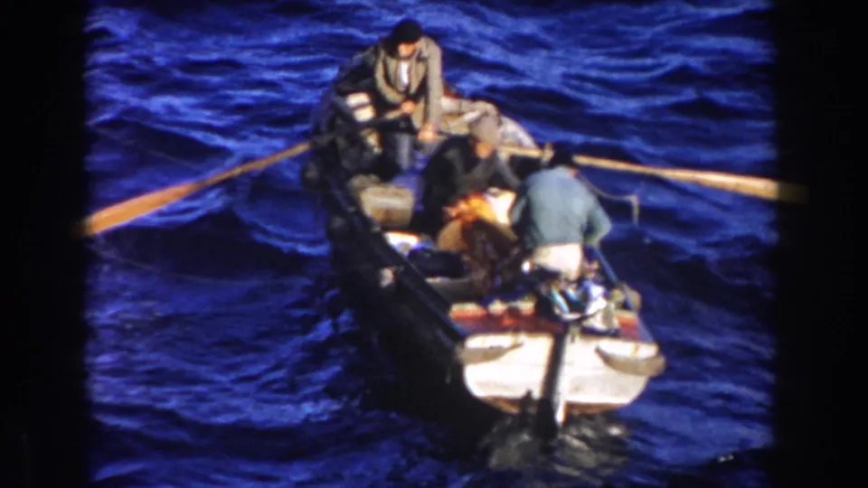 The rowboat angles through choppy water in Gibraltar, with one man standing near the stern and two men seated among gear.