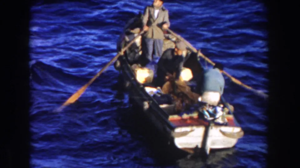 A high-angle 8mm view shows three men in a small rowboat on deep blue water, with oars spread and gear clustered near the stern.