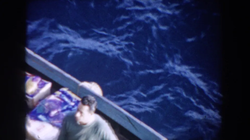 1959 Gibraltar frame with an adult man in a green shirt standing or bracing inside a small white boat on choppy sea.