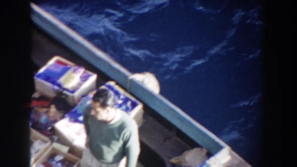 1959 Gibraltar frame showing an adult in a small boat near packed supplies, filmed from above with deep blue water in the background.
