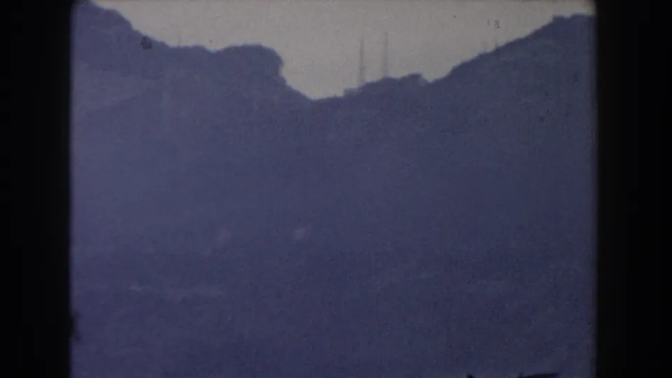 1959 Gibraltar 8mm frame showing a dark hazy hillside and ridge, with pale sky, soft focus, grain, and small film blemishes.