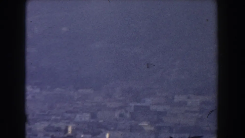 Soft, grainy 1959 Gibraltar aerial view with clustered coastal buildings beneath a hazy blue-grey sky.