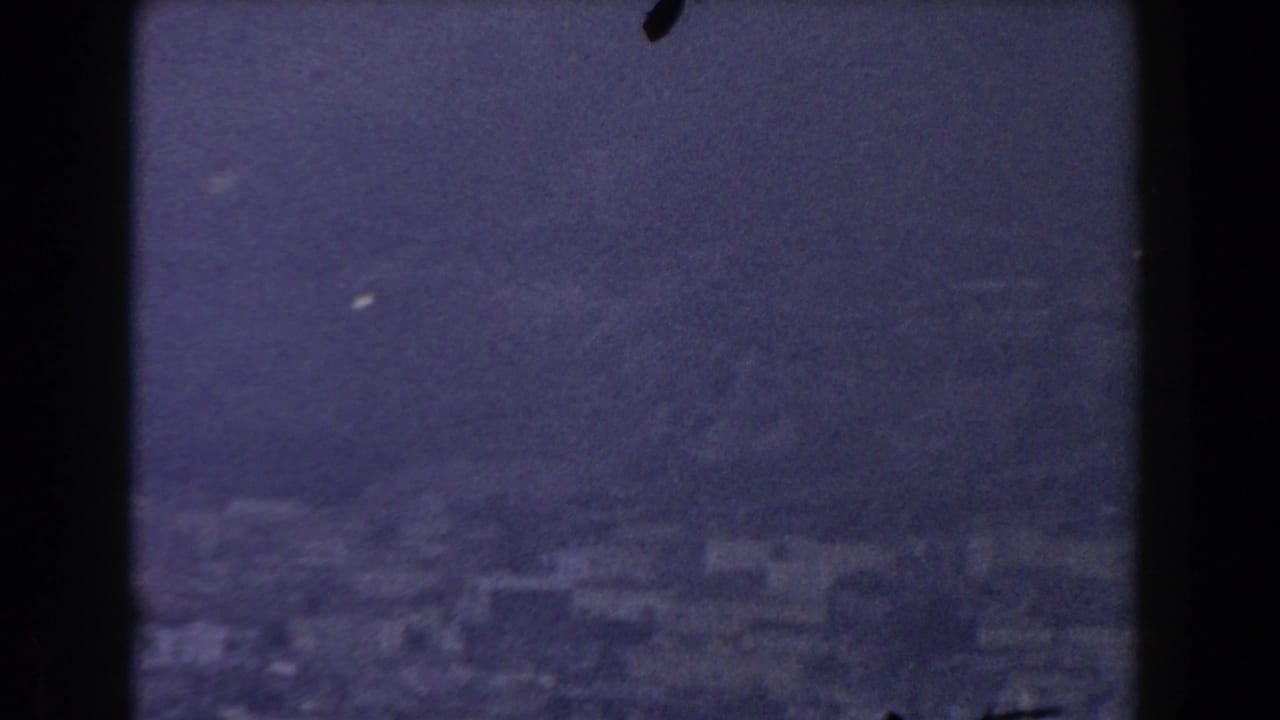 1959 Gibraltar home-movie view of a hazy shoreline with pale buildings below steep mountain terrain across the water.