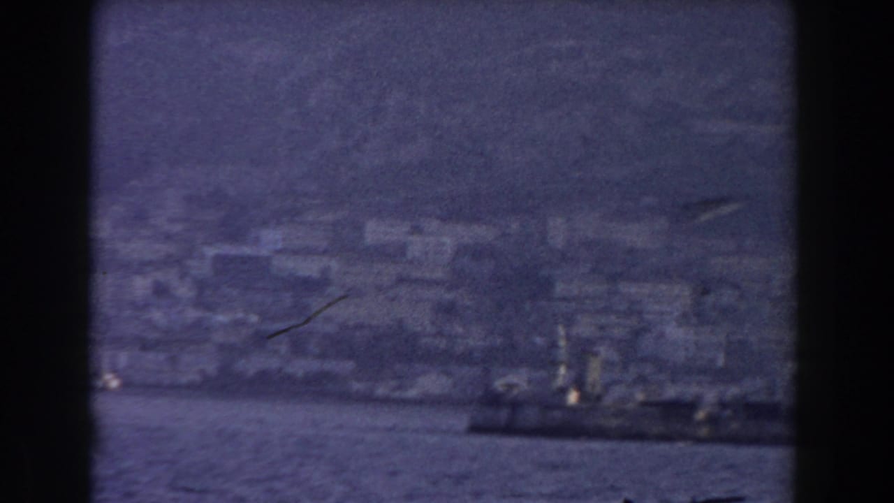 A dark vessel passes near Gibraltar's hazy waterfront, with pale buildings and steep terrain in the background.