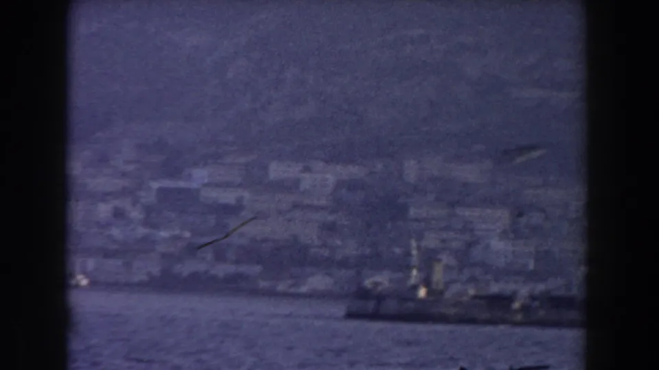 A dark vessel passes near Gibraltar's hazy waterfront, with pale buildings and steep terrain in the background.