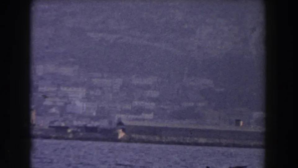 A long dark ship crosses the foreground of a 1959 Gibraltar harbor view, backed by blurred shoreline buildings and mountain haze.